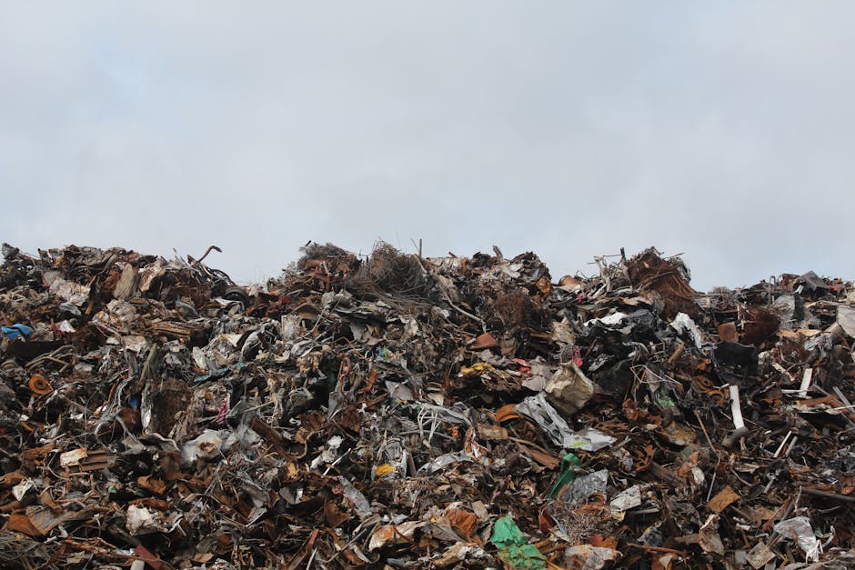 A large mound of mixed waste materials dominates the foreground, consisting of broken pieces of wood, shredded paper, plastic debris, metal scraps, and tangled textiles, all piled haphazardly on a paved surface. The textures vary from rough and jagged wood fragments to smooth, crumpled plastic and shiny metal objects. The color palette includes dark browns, faded whites, vibrant greens, and occasional bright hues from packaging and plastic items. The waste appears to have been accumulated over time, with some materials compressed or partially buried beneath others, creating a chaotic, unorganized heap. In the background, an overcast sky with grey clouds contributes to a subdued atmosphere, highlighting the industrial or landfill setting. This scene illustrates the volume of rubbish that waste disposal services such as Waste Disposal Kensington manage, often requiring specialized equipment for off-site or private collection efforts, rather than relying solely on local authority rubbish collection. The image underscores the importance of professional waste removal in maintaining environmental standards and preventing illegal dumping near spaces like Kensington Gardens.