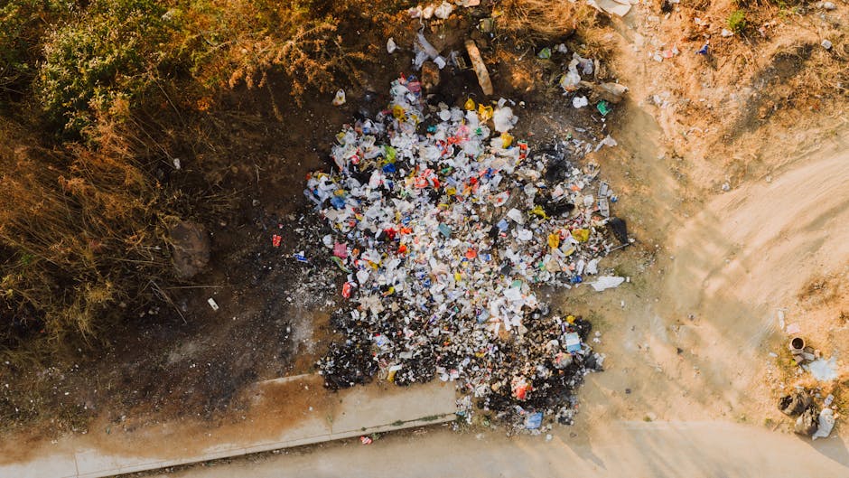 An aerial view showing a large pile of mixed household waste and plastic rubbish piled against a patch of bare, uneven soil and surrounding dry, shrub-covered terrain. The pile includes various types of plastic containers, wrappers, and discarded packaging, with some debris scattered on the ground nearby. To the right of the rubbish, a concrete sidewalk runs adjacent to the area, with a small metal pipe embedded in the ground at the edge of the pavement. The environment appears to be an outdoor space with natural lighting, highlighting the contrast between the dark, burnt-looking soil and the colorful debris. This scene illustrates an instance of private waste accumulation that could benefit from professional rubbish removal services, such as those provided by Waste Disposal Kensington, to ensure proper clearance and adherence to local waste management regulations.