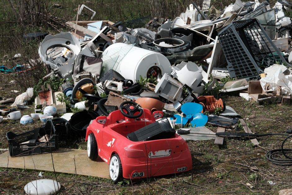 A large mound of mixed waste materials dominates the foreground, consisting of broken pieces of wood, shredded paper, plastic debris, metal scraps, and tangled textiles, all piled haphazardly on a paved surface. The textures vary from rough and jagged wood fragments to smooth, crumpled plastic and shiny metal objects. The color palette includes dark browns, faded whites, vibrant greens, and occasional bright hues from packaging and plastic items. The waste appears to have been accumulated over time, with some materials compressed or partially buried beneath others, creating a chaotic, unorganized heap. In the background, an overcast sky with grey clouds contributes to a subdued atmosphere, highlighting the industrial or landfill setting. This scene illustrates the volume of rubbish that waste disposal services such as Waste Disposal Kensington manage, often requiring specialized equipment for off-site or private collection efforts, rather than relying solely on local authority rubbish collection. The image underscores the importance of professional waste removal in maintaining environmental standards and preventing illegal dumping near spaces like Kensington Gardens.