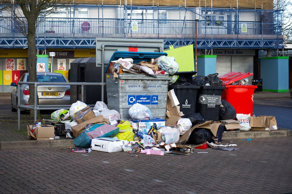 A large collection of overflowing rubbish and waste materials stacked around and inside several open-top communal waste bins situated on a paved parking area. The waste includes various cardboard boxes, paper and plastic bags, crumpled packaging, and scattered trash spilling onto the surrounding ground. Some of the waste is tied in plastic bags, while other items are loose, creating a cluttered scene. Behind the bins, there is a metal railing separating the rubbish area from parked vehicles, with a grey car partially visible to the left. In the background, a building with a blue metal scaffolding structure and a balcony is visible, along with storefronts that display signage, indicating a commercial or retail street environment. The scene appears to be outdoors, under natural daylight, with a tree on the left side adding to the urban setting. This image illustrates issues related to private waste disposal and the importance of proper rubbish removal services, such as those provided by Waste Disposal Kensington, to manage such waste effectively.
