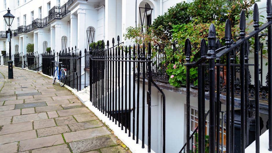 A row of four wheelie bins positioned along a residential street pavement next to a tall, dense hedge. The trash bins include three black bins with orange lids, one blue bin, and a green-lidded bin, all marked with the RMN logo. They are situated near a black decorative lamppost and a wooden fence, with autumn leaves scattered across the concrete sidewalk and curb. The street curves gently into the distance, lined with trees bearing autumn foliage, and a parked silver car is visible further along the road. The scene depicts typical urban waste disposal arrangements, which may involve private rubbish collection services like Waste Disposal Kensington, offering an alternative to traditional local authority collection for residents seeking on-site or independent rubbish removal solutions.