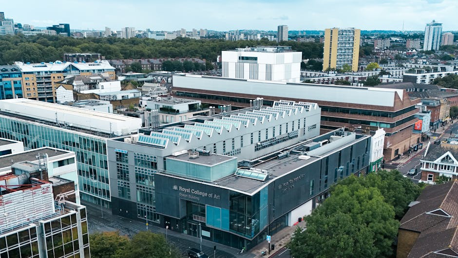 An aerial view of an urban area featuring a large, modern building with a prominent sign reading 'Royal College of Art,' constructed with dark grey and glass facade elements, situated among other commercial and residential structures with varying architectural styles. The building's flat roof is equipped with skylights and mechanical equipment, and nearby is a mix of modern glass-fronted offices and older brick buildings. Surrounding the central structure are streets with some vehicles and a street-level view of trees filtering sunlight onto sidewalks. In the background, a densely built cityscape extends with mid-rise and high-rise buildings, interspersed with patches of green spaces. The environment appears well-maintained, illustrating a typical city centre scene which could be associated with independent waste collection or on-site clearing services provided by waste disposal specialists in London, such as Waste Disposal Kensington, supporting effective rubbish removal and private waste handling amidst urban development.
