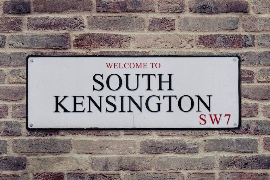 A rectangular metal street sign mounted on a textured red brick wall, displaying the words 'WELCOME TO SOUTH KENSINGTON' in bold black capital letters, with 'SW7' in smaller red letters at the bottom right corner. The sign has a white background with rounded corners and is attached with four visible screws at each corner. The brick wall behind the sign features horizontal rows of bricks in various shades of reddish-brown, with some mortar joints showing signs of weathering. The lighting appears natural and even, highlighting the contrast between the crisp, clean sign and the aged, rough surface of the brick wall, representing a typical urban setting that could relate to private or alternative waste disposal options in a residential or commercial area.