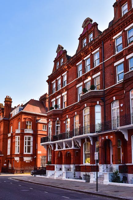 A tall, red-brick Victorian-style residential building situated on a city street with a clear blue sky overhead. The structure features multiple floors with large sash windows, some with small balconies or flower pots on window sills. The building has decorative elements such as white trim, arched window surrounds, and a distinctive gabled roof with dormer windows. The street-level entrance includes a set of stairs leading up to a covered porch area supported by white decorative brackets. To the left, similar red-brick buildings are visible, also with bay windows and detailed masonry. The scene appears to be in an urban area aligned with private property, which might be part of an independent collection or on-site clearance, consistent with services offered by Waste Disposal Kensington. The environment is well-maintained with a parked car and a street sign in the foreground, and minimal pedestrian activity, emphasizing a neat, residential setting typical in Kensington and Chelsea.