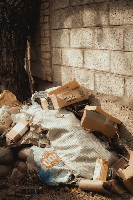 A pile of mixed waste materials is gathered against a rough brick wall, situated on a dirt surface outdoors. The collection includes flattened cardboard boxes, some with printed logos and barcodes, and crumpled brown paper bags. A large, textured, woven bag or sack in a off-white or light grey color is partially visible, serving to contain or support the waste. The cardboard boxes vary in size and are slightly torn, with some leaning against each other or lying flat. The environment appears to be outdoors, possibly an alleyway or side yard, with natural daylight illuminating the scene. A dark tree trunk is partially visible on the far left side, and the brick wall behind the waste is unfinished or weathered, with mortar joints clearly visible. The scene reflects typical private rubbish collection efforts, consistent with services provided by Waste Disposal Kensington for on-site waste clearance and alternative rubbish removal solutions.