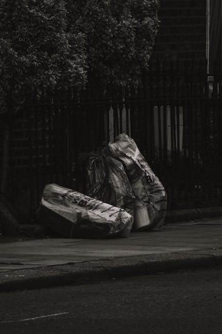 A pile of mixed waste materials is gathered against a rough brick wall, situated on a dirt surface outdoors. The collection includes flattened cardboard boxes, some with printed logos and barcodes, and crumpled brown paper bags. A large, textured, woven bag or sack in a off-white or light grey color is partially visible, serving to contain or support the waste. The cardboard boxes vary in size and are slightly torn, with some leaning against each other or lying flat. The environment appears to be outdoors, possibly an alleyway or side yard, with natural daylight illuminating the scene. A dark tree trunk is partially visible on the far left side, and the brick wall behind the waste is unfinished or weathered, with mortar joints clearly visible. The scene reflects typical private rubbish collection efforts, consistent with services provided by Waste Disposal Kensington for on-site waste clearance and alternative rubbish removal solutions.
