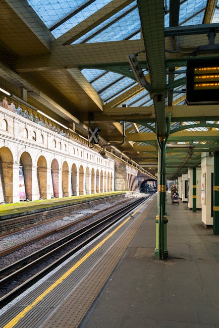A view of a train station platform with multiple railway tracks extending into the distance, set beneath a large, curved glass and metal canopy with a yellow underside. The platform features a textured yellow tactile strip along the edge and a grey paved surface. On the right side, green metal support poles and canopy framework dominate the scene, with a few passengers visible in the background, some seated on benches, and a small cart or luggage trolley. Along the opposite side of the tracks, a brick wall with arched openings and faint advertisements is visible, indicating an historic building facade integrated into the station environment. The lighting is natural, suggesting daytime, and the overall scene emphasizes urban public transport infrastructure, with a subtle connection to the context of managed or alternative waste handling within public spaces, such as rubbish clearance or station management, which companies like Waste Disposal Kensington might support through service offerings for station maintenance or private waste collection.