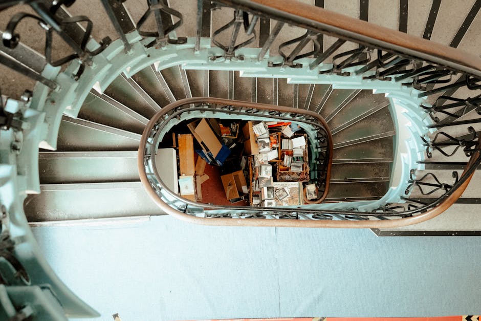 A view looking down a spiral staircase with metal railings and a wooden handrail, revealing a cluttered collection of various discarded cardboard boxes, some flattened and others partially assembled, along with miscellaneous packing materials and small household items scattered on the floor at the bottom of the staircase. The staircase has a light teal painted central column and black metal balusters, with natural lighting illuminating both the steps and the debris below. The environment appears to be an interior space, possibly a staircase in a residential building or communal area, which is temporarily used for stacking rubbish awaiting removal. The image relates to waste management and rubbish removal services, such as those offered by Waste Disposal Kensington, especially in the context of private or alternative collection options for properties with limited access or tight stairways, where on-site clearance might be required.