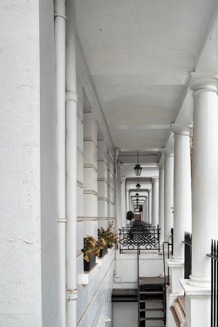 A long, narrow view of a white, enclosed balcony or corridor featuring a series of classical-style columns supporting an overhead ceiling. The columns are cylindrical with smooth finishes, painted white, and evenly spaced along the length of the space. On the left side, there are white-painted walls with decorative molding, small black wrought iron fences, and planters filled with brownish-orange flowers. The floor is visible in the distance, and there are outdoor-style lanterns hanging from the ceiling, providing ambient lighting. The environment appears clean and well-maintained, with natural light illuminating the scene. This type of setting could be associated with private residential buildings or property with multiple units, where independent or on-site waste collection might be relevant, aligning with alternative waste handling services. Waste Disposal Kensington may support such locations seeking efficient rubbish removal for communal or private exterior spaces, highlighting the importance of proper waste management in areas with architectural detailing and outdoor features.