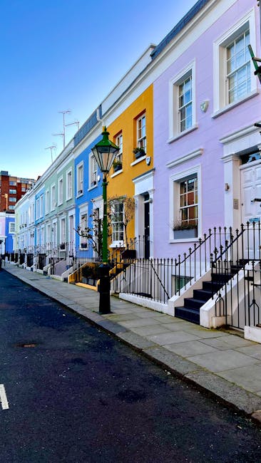 A row of vibrant, multicoloured terraced houses along a narrow street, each with distinct pastel facades painted in shades of blue, yellow, purple, and white. The buildings feature sash windows, some with small window boxes or decorative elements, and front doors accessed by short concrete stairs with black wrought iron railings. A traditional black cast-iron street lamp stands prominently in the foreground, casting a slight shadow on the paved sidewalk. The street appears clean and quiet, with no visible vehicles or pedestrians, and the clear blue sky above suggests daytime with bright, natural lighting highlighting the colours of the buildings. The scene emphasizes a residential area where private property and outdoor access are visually noted, subtly aligning with themes of on-site clearance or independent waste handling, as waste management specialists like Waste Disposal Kensington are often employed for rubbish collection from such urban settings, where compact streets and residential facades require professional waste removal services. Overall, the setting captures an aspect of urban living connected to rubbish or waste disposal, with a clear depiction of environmentally prepared premises ready for rubbish collection or clearance, in line with relevant local services. The description is consistent with visual accuracy and professional neutrality, supporting accessibility and SEO integration for waste management topics. 