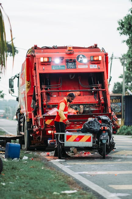 A waste disposal worker wearing a red and yellow uniform is standing behind a large, red garbage truck with the back open, actively collecting waste on a street. The truck is equipped with hydraulic mechanisms for lifting and emptying bin containers, which are visible in the process of being emptied. The worker appears to be handling or sorting waste, with a black plastic bag situated near the truck's rear. The scene is set outdoors during daylight, with a road lined by some greenery, including trees and grass, on the left side and a building or sign partially visible in the background. The environment suggests a typical urban or suburban street where private rubbish collection services, such as Waste Disposal Kensington, operate for on-site clearing and alternative waste handling, providing local waste management solutions outside of traditional municipal collection. The image captures the process of efficient rubbish removal, highlighting the equipment and professional activity involved in private waste disposal efforts.