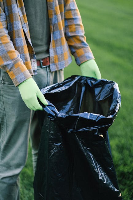 A person wearing a plaid shirt with yellow, gray, and blue checks, along with green gloves, is holding open a large black rubbish bag made of glossy plastic material. The bag is partially filled and positioned upright, ready for waste disposal. The individual is standing outdoors on a grassy area, with a blurred background of green grass and foliage, suggesting a park or garden setting. The scene emphasizes waste collection or rubbish removal activity, illustrating a private or independent approach to disposal that Waste Disposal Kensington might support through their rubbish removal services. The natural lighting highlights the textures of the gloves, fabric, and the shiny surface of the rubbish bag, creating a clear, professional visual scene focused on waste handling. The overall composition aligns with themes of on-site clearance and waste management, matching the context of alternative rubbish collection options in Kensington.