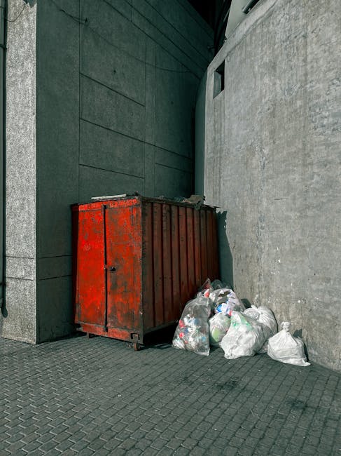 A waste disposal worker wearing a red and yellow uniform is standing behind a large, red garbage truck with the back open, actively collecting waste on a street. The truck is equipped with hydraulic mechanisms for lifting and emptying bin containers, which are visible in the process of being emptied. The worker appears to be handling or sorting waste, with a black plastic bag situated near the truck's rear. The scene is set outdoors during daylight, with a road lined by some greenery, including trees and grass, on the left side and a building or sign partially visible in the background. The environment suggests a typical urban or suburban street where private rubbish collection services, such as Waste Disposal Kensington, operate for on-site clearing and alternative waste handling, providing local waste management solutions outside of traditional municipal collection. The image captures the process of efficient rubbish removal, highlighting the equipment and professional activity involved in private waste disposal efforts.