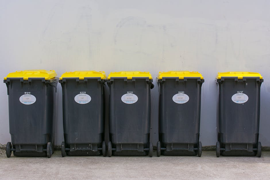 Five large wheeled rubbish bins are lined up neatly on a paved surface against a plain, light-colored wall. The bins are made of dark grey plastic with a matte finish and have bright yellow lids that are securely closed. Each bin features a small oval white label with black text, affixed near the top front, providing identification or waste type information. The bins are positioned side by side, with minimal space between them, and appear to be in an outdoor setting, possibly a driveway or loading area. The environment is well-lit with natural light, casting subtle shadows on the ground and on the wall behind. The scene, part of an organized waste collection area, subtly reflects the services of private waste disposal or independent rubbish collection, such as those offered by Waste Disposal Kensington, emphasizing efficient on-site clearance and waste management for residential or commercial premises.