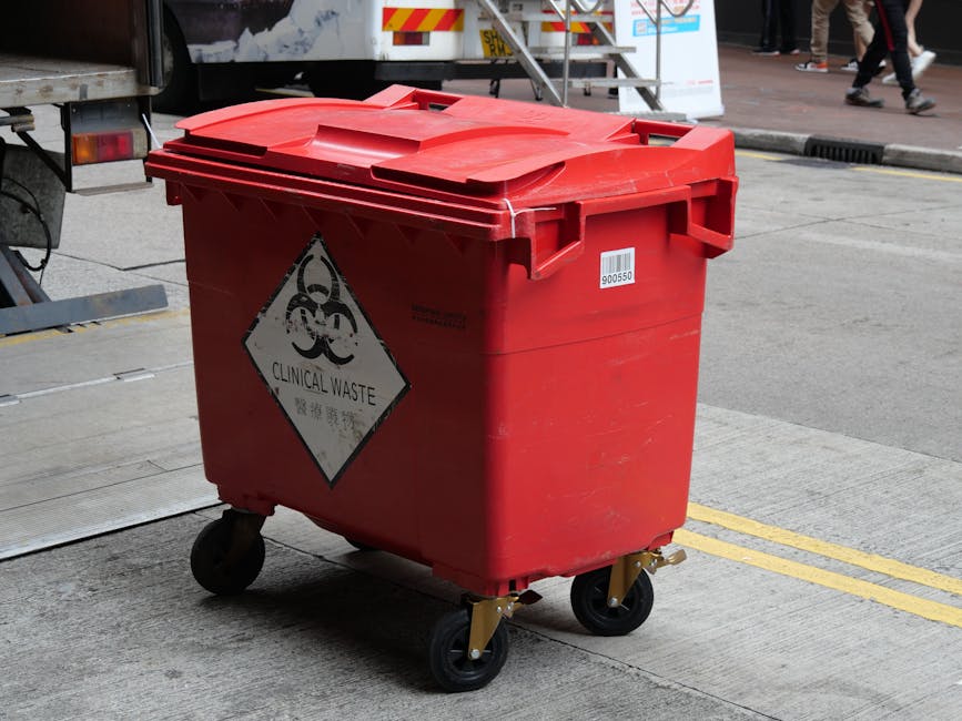 A red plastic bin with a hinged lid, labeled with a biohazard warning and the words 'Clinical Waste' on a diamond-shaped sign. The bin is mounted on four black caster wheels with visible orange metal brackets, situated on a concrete sidewalk next to a curb in an urban environment. In the background, a parked truck with reflective yellow and red markings is partially visible, along with a metal staircase and pedestrians walking nearby on the street. The scene appears to be outdoors during daylight, with natural lighting casting soft shadows, highlighting the bin's textured surface and the surrounding pavement. This setup reflects a typical container used for the collection and temporary storage of clinical or medical waste, aligning with waste management services that handle hazardous or specialized rubbish removal in urban areas, such as those provided by Waste Disposal Kensington.