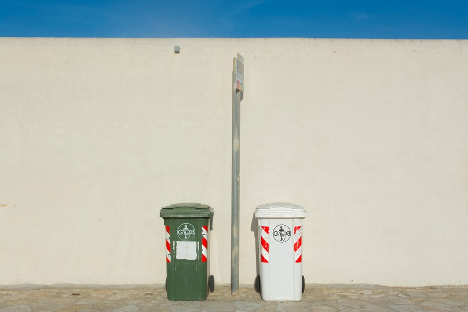 Five large wheeled rubbish bins are lined up neatly on a paved surface against a plain, light-colored wall. The bins are made of dark grey plastic with a matte finish and have bright yellow lids that are securely closed. Each bin features a small oval white label with black text, affixed near the top front, providing identification or waste type information. The bins are positioned side by side, with minimal space between them, and appear to be in an outdoor setting, possibly a driveway or loading area. The environment is well-lit with natural light, casting subtle shadows on the ground and on the wall behind. The scene, part of an organized waste collection area, subtly reflects the services of private waste disposal or independent rubbish collection, such as those offered by Waste Disposal Kensington, emphasizing efficient on-site clearance and waste management for residential or commercial premises.