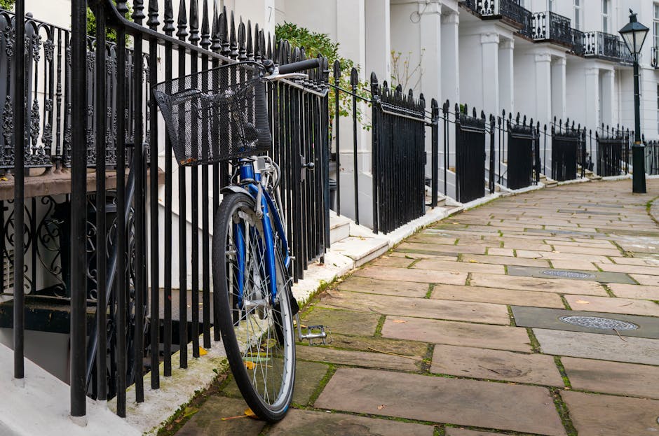 The image depicts a narrow street or sidewalk lined with white terraced houses featuring black wrought iron railings along the front. Positioned against one of these railings is a blue bicycle with a black front basket, leaning on its kickstand. The bicycle has a metallic frame with a textured black seat and standard black tires. The paving consists of large, rectangular stone slabs with visible grout lines, some patches of moss or dirt in the joints, and a few fallen leaves scattered across the surface. In the background, the facades of the houses display classic architectural elements such as white columns supporting small balconies with ornate black railings, and a wall-mounted lantern on the rightmost building. The environment appears to be well-maintained, with a clean appearance and natural daylight illuminating the scene. The setting suggests an urban residential area where alternative waste handling or private rubbish collection services, such as those provided by Waste Disposal Kensington, might operate to assist with rubbish removal from properties behind the railings.