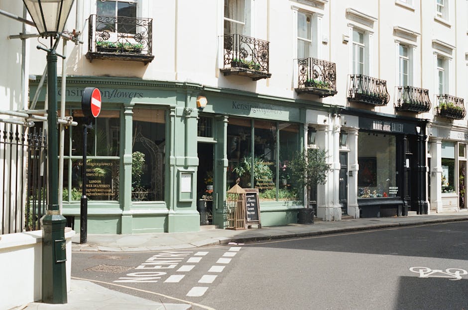 A street-level view of a row of multi-storey buildings with retail storefronts. The first shop has a light green facade with large glass windows displaying plants and flowers inside, and a small black chalkboard sign outside advertising floral arrangements. The building features decorative white trim around the windows and a narrow sidewalk in front. To the left, a black metal street lamp and a pole with a red and white street sign are visible. The background includes additional upper-story windows with wrought-iron balcony railings, some with potted plants. The street surface has painted markings, including a bicycle lane symbol and a crosswalk pattern. The scene is illuminated by natural daylight, highlighting the textures of the building finishes and the street environment, fitting the context of a location where residents or businesses might arrange for private waste collection or rubbish removal services, as offered by Waste Disposal Kensington.